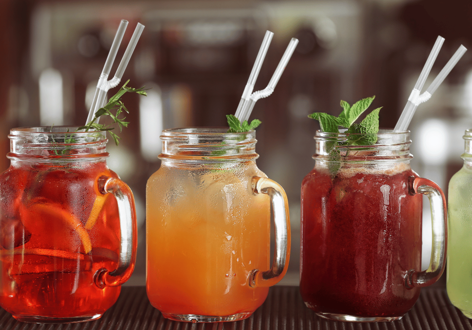 Three mason jar drinks with ice and mint on a bar counter, left red, middle orange, right dark red.